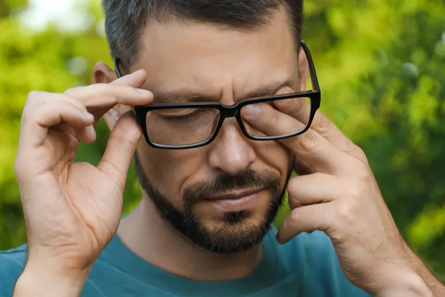 A man with a beard and glasses standing outside and wearing a blue t-shirt lifts his glasses and rubs his eyes due to allergy irritation. Get treatment for allergic conjunctivitis from Dr. Bahar Amin in North York.
