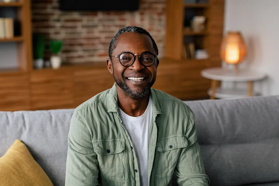 A middle aged black man in a green button up shirt sits on his couch smiling, happy with his treatment for andropause and low testosterone from Dr. Bahar Amin in North York.
