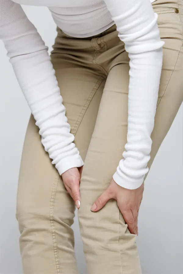 A woman wearing a white long-sleeve shirt and tan pants, clutching her knee, suffering from arthritis. Get arthritis treatment from Dr. Bahar Amin in North York.