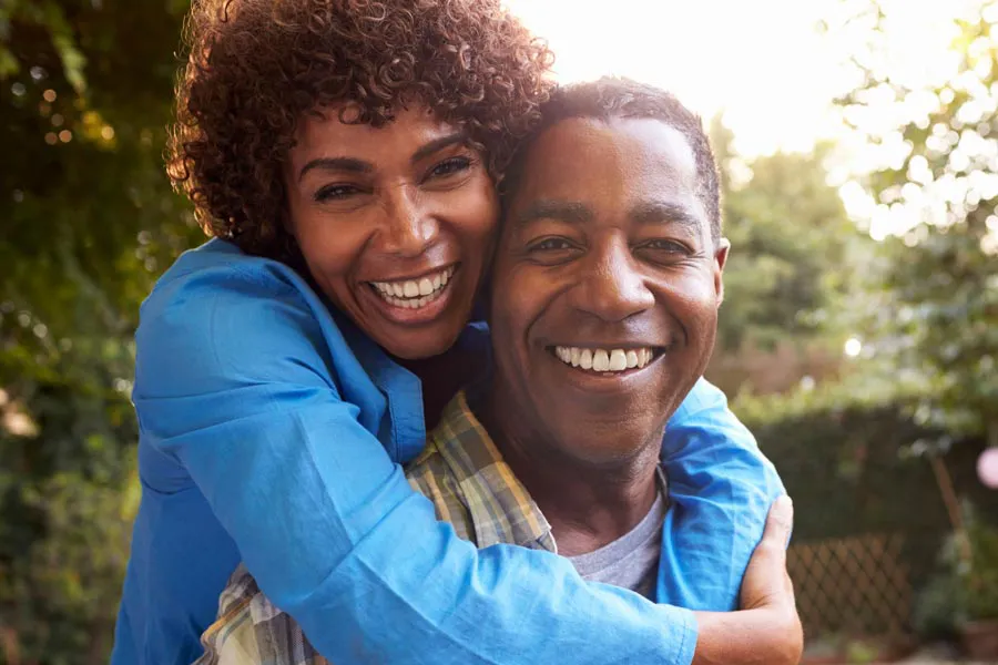 A middle-aged African American couple outside, the woman embracing the man from behind pressing her cheek to his, both are smiling due to successful Autoimmune Disorder treatment from Dr. Bahar Amin in North York.