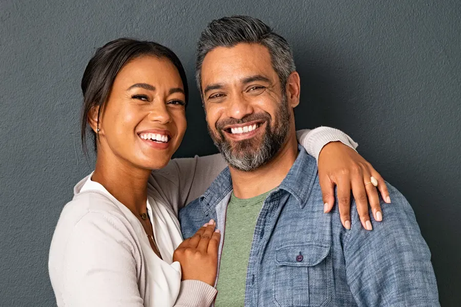 A middle-aged couple with their arms around each other standing against a gray background and smiling, happy with the bioidentical hormone therapy they received from Dr. Bahar Amin in North York.