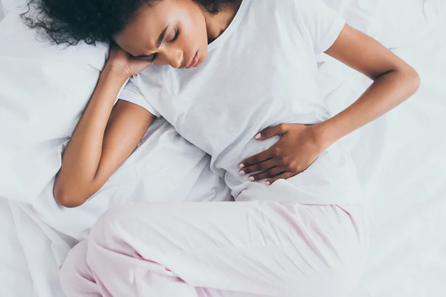 A dark-skinned woman in white clothes lays on her bed clutching her middle before getting treatment for Crohn's Disease from Dr. Bahar Amin in North York.
