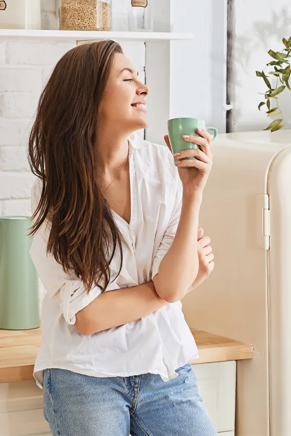 A woman with a white blouse an jeans drinking tea in her kitchen, smiling after getting treatment for Crohn's Disease from Dr. Bahar Amin in North York.