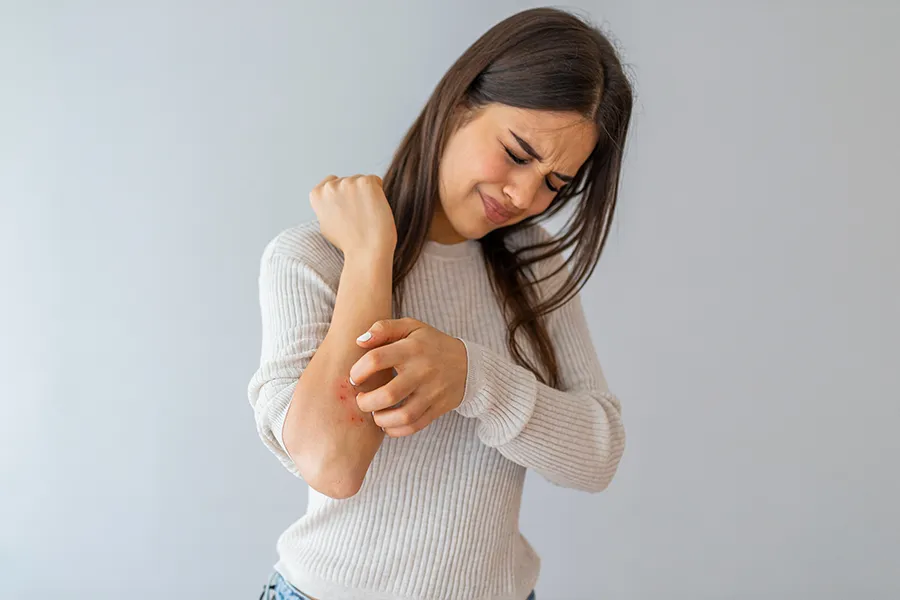A young brunette woman scratches her forearm and winces in discomfort. Get treatment for eczema from Dr. Bahar Amin in North York.