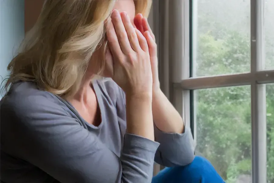 A blonde woman in a gray shirt sits in front of a window on a rainy day covering her eyes with her hands. Get treatment for Grave's Disease from Dr. Bahar Amin in North York.