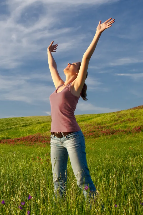 A woman in a tank top and jeans looks up with her arms outstretched to the sky in a field after successful treatment for Grave's Disease from Dr. Bahar Amin in North York.