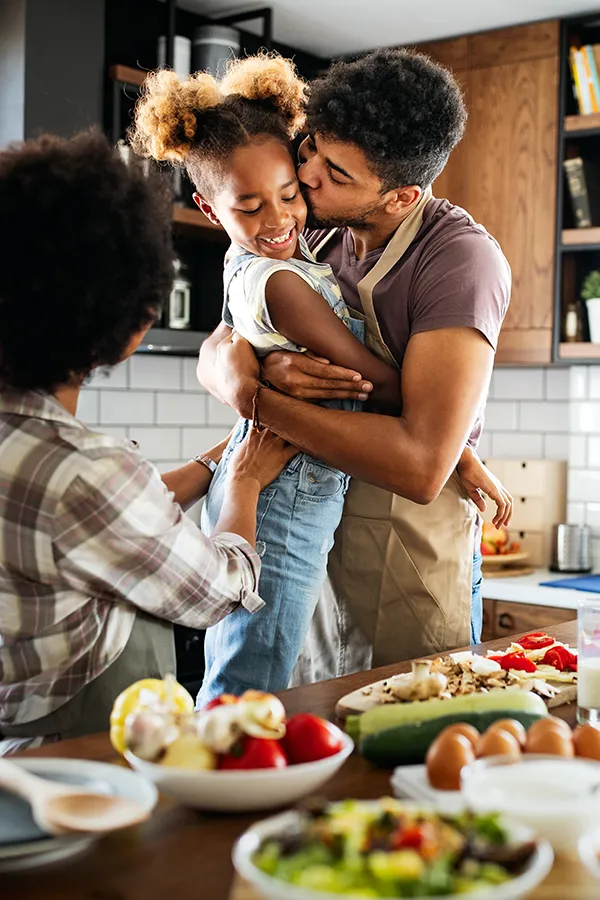A man holds his daughter and kisses her cheek as the family makes a healthy dinner in the kitchen. Get help for digestive issues and gut health from Dr. Bahar Amin in North York.