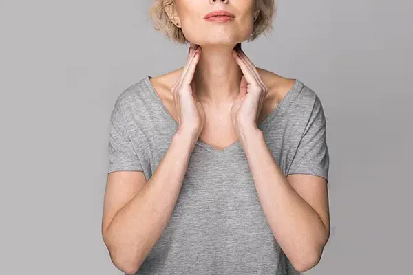 An older blonde woman in a gray t-shirt holds her hands to her neck checking for signs of thyroid problems. Get help for Hashimoto's Thyroiditis from Dr. Bahar Amin in North York.