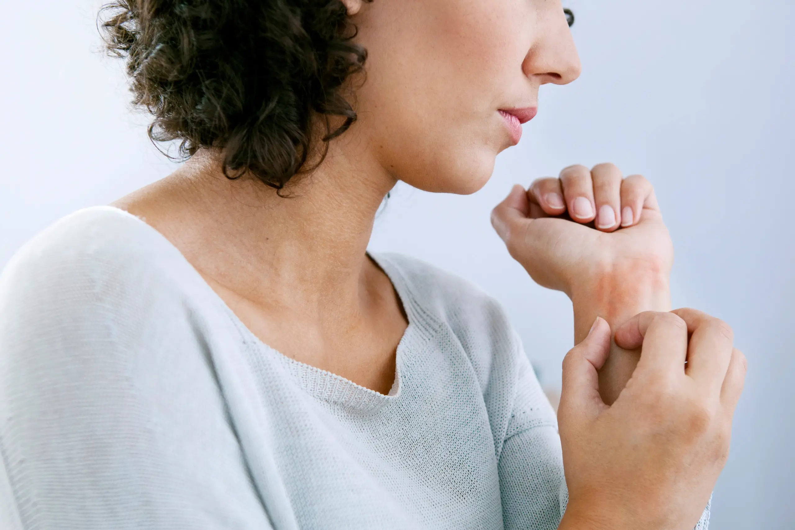 A woman with curly brown hair and a light gray shirt scratches her wrist due to hives. Get treatment for hives from Dr. Bahar Amin in North York.