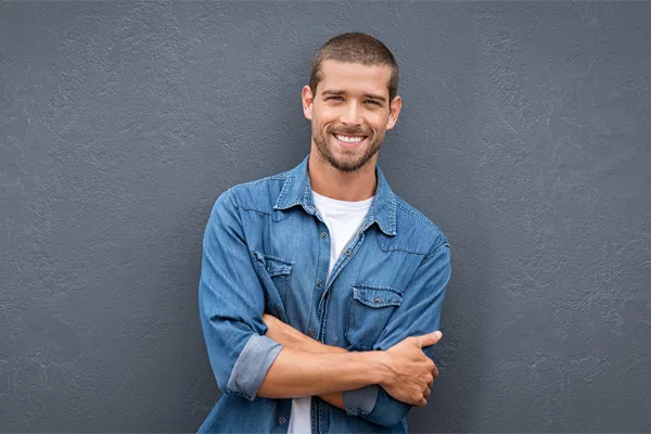 A man in a denim shirt stands smiling against a gray-blue wall, pleased with his testosterone hormone therapy from Dr. Bahar Amin in North York.