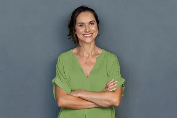 A woman in a green shirt stands smiling against a gray-blue wall, pleased with her hormone therapy from Dr. Bahar Amin in North York.