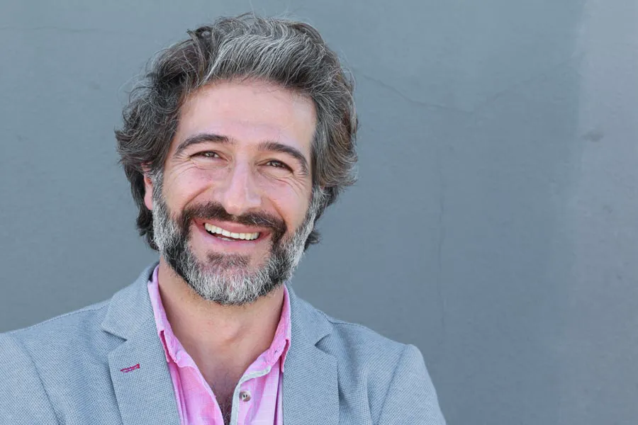 A smiling man with salt and pepper hair and beard wearing a pink shirt and gray blazer stands agains a gray wall. Get hypogonadism treatment from Dr. Bahar Amin in North York.