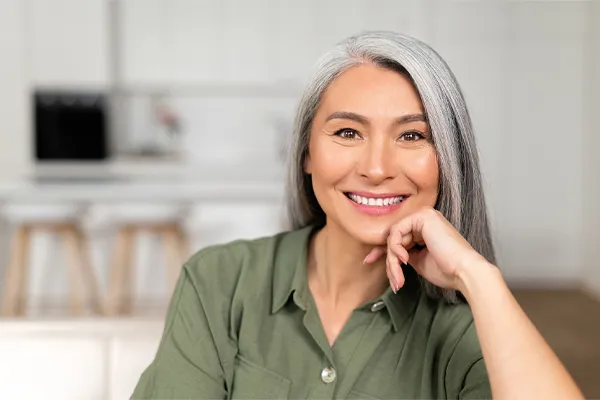 An asian woman with long gray hair and a green shirt sitting and smiling on her couch, happy with her menopause care from Dr. Bahar Amin in North York.