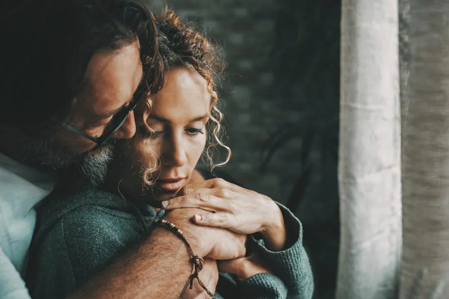 A man embraces a woman from behind in a dimly lit room, their somber expressions showing concerns about a neurodegenerative disease they will get treated by Dr. Bahar Amin in North York.