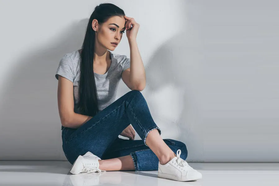 A young brunette woman in a gray t-shirt and jeans sits against a gray wall with her head resting on her hand and a distressed expression. Get help for Polycystic Ovarian Syndrome (PCOS) from Dr. Bahar Amin in North York.