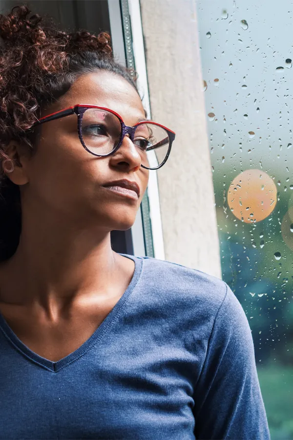 A black woman with with a somber expression and glasses and a gray shirt stares out the window on a rainy day. Get treatment for Premenstrual Dysphoric Disorder from Dr. Bahar Amin in North York.