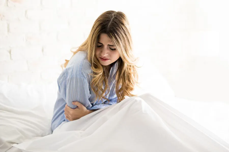 A woman with long blonde hair sits in her bed holding her stomach in pain, considering getting care for PMS from Dr. Bahar Amin in North York.