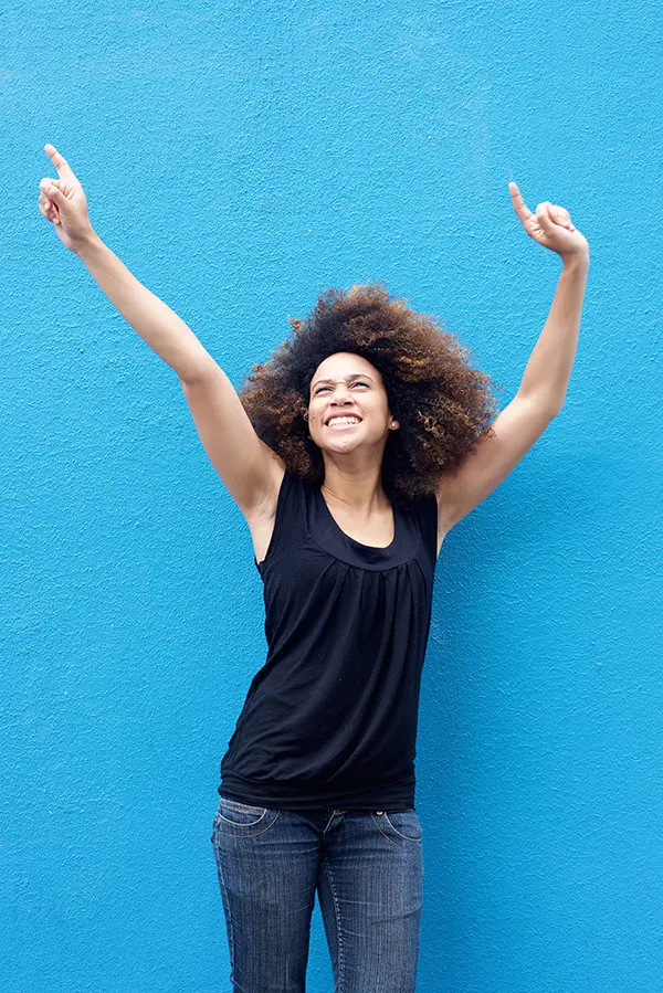 A woman in a dark blue tank top standing in front of a bright blue wall, raising her arms in celebration of relief from PMS from Dr. Bahar Amin in North York.