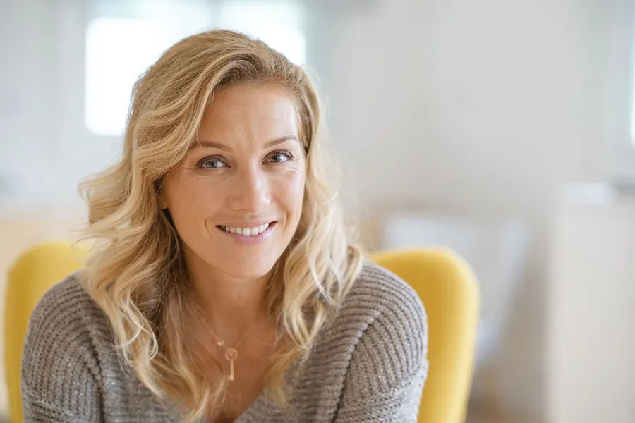 A blonde woman wearing a a gray sweater sits inside in a yellow chair, smiling and happy with her perimenopause treatment from Dr. Bahar Amin in North York.