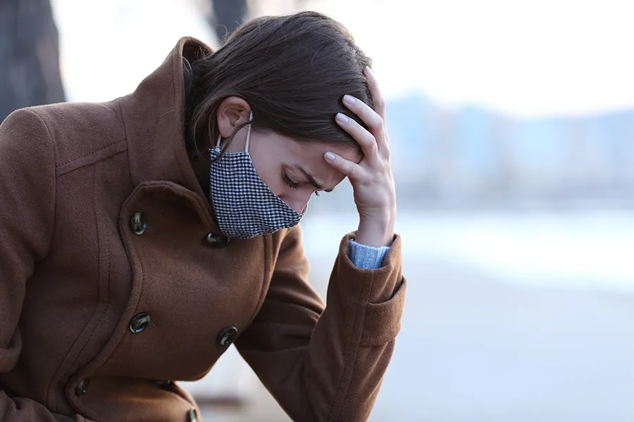 A woman in a mask sits outside with her hand on her forehead in pain. Get long covid treatment from Dr. Bahar Amin in North York.