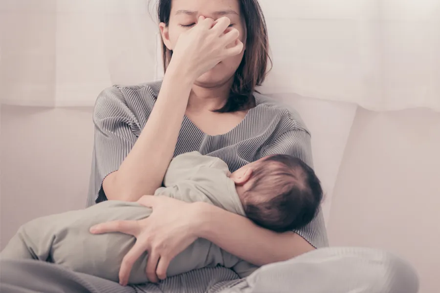 A woman nursing an infant on couch rubs her eyes with exhaustion. Get treatment for postpartum hormone imbalance from Dr. Bahar Amin in North York.
