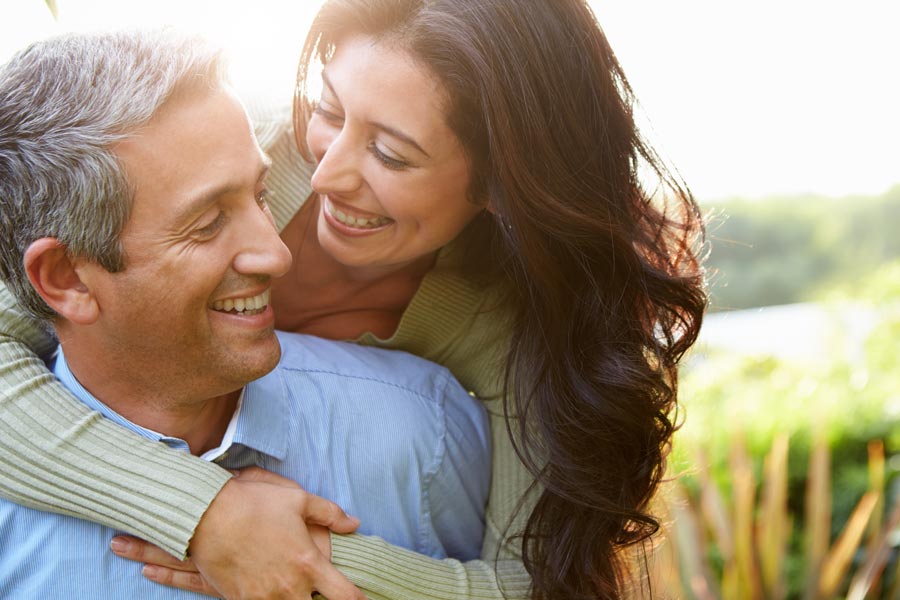 Smiling middle-aged couple embracing outdoors in a sunlit natural setting. Receive personalized care through Dr. Bahar Amin's metabolic and hormone reset program offered in North York.